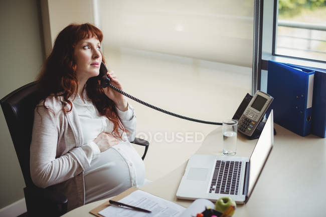 Беременная деловая женщина трогает живот, разговаривая по телефону в офисе — стоковое фото