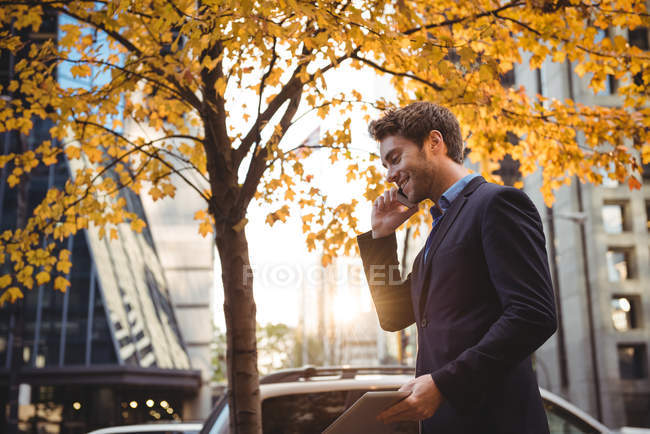 Усміхнений бізнесмен розмовляє на мобільному телефоні і тримає цифровий планшет на вулиці — стокове фото