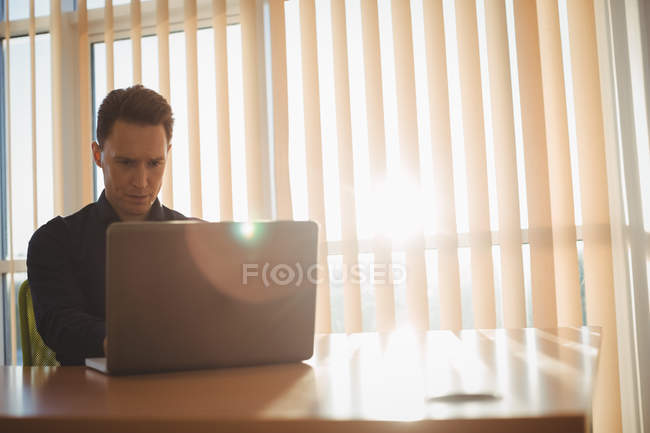 Executivo masculino usando laptop perto de persianas no escritório — Fotografia de Stock