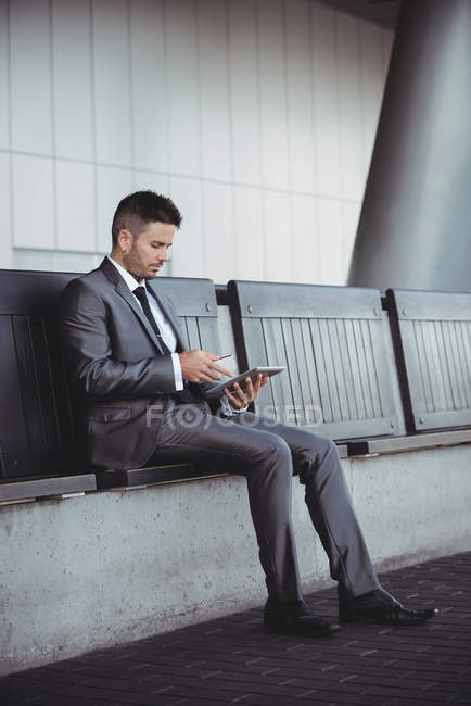 Бизнесмен использует цифровой планшет, сидя на скамейке в офисном кампусе — стоковое фото