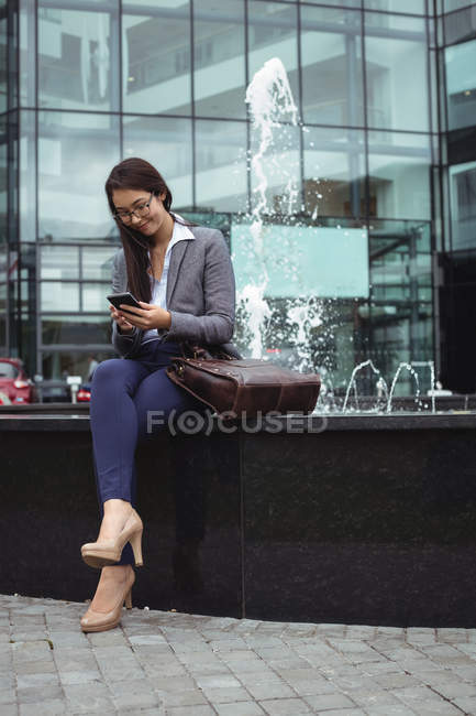Mujer de negocios sentada cerca de la fuente y usando teléfono móvil fuera del edificio de oficinas - foto de stock