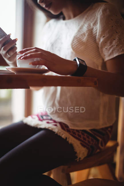 Mulher usando telefone celular enquanto toma uma xícara de café no café — Fotografia de Stock