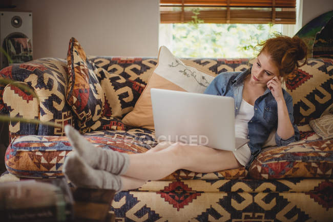 Nachdenkliche Frau benutzt Laptop auf Sofa zu Hause — Stockfoto