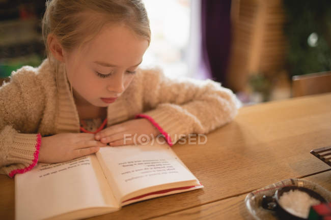 Chica rubia sentada en la mesa y leyendo libro en casa - foto de stock