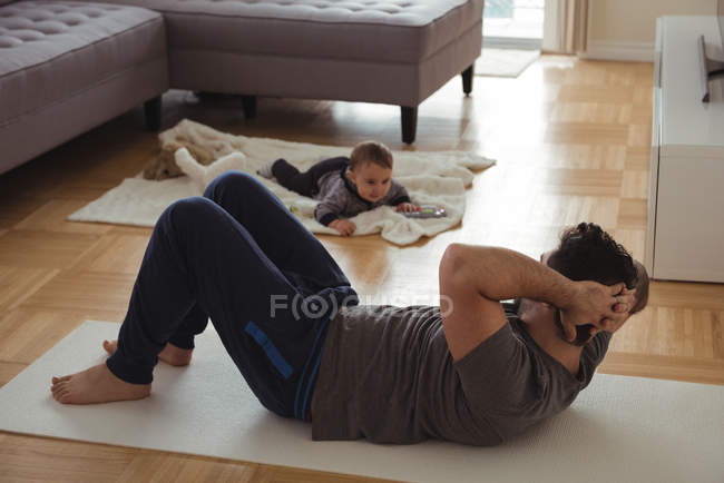 Padre haciendo ejercicio mientras el bebé juega en segundo plano en casa - foto de stock