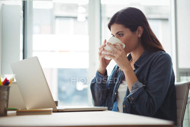 Business executive having cup of coffee while using laptop in office — Stock Photo