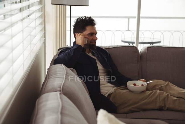 Homem falando no celular enquanto toma café da manhã no sofá — Fotografia de Stock