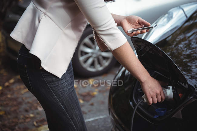 Seção média da mulher usando telefone celular ao carregar carro elétrico na rua — Fotografia de Stock