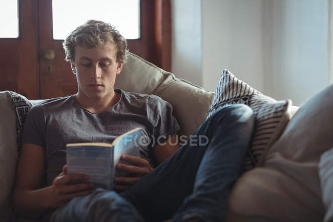 Hombre sentado en sofá leyendo un libro en la sala de estar - foto de stock