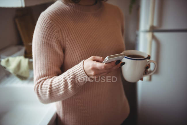 Frau benutzt Handy, während sie zu Hause in der Küche Kaffee trinkt — Stockfoto