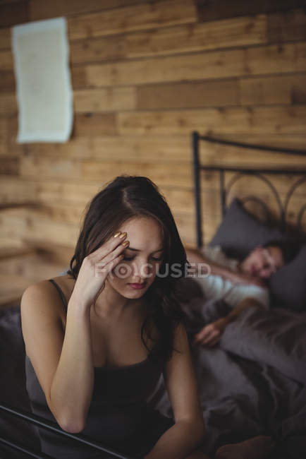 Aufgebrachte Frau sitzt auf einem Bett, während Mann im Hintergrund im Schlafzimmer schläft — Stockfoto
