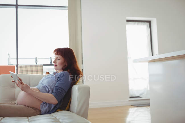 Pregnant woman digital tablet while relaxing on sofa in living room at home — Stock Photo