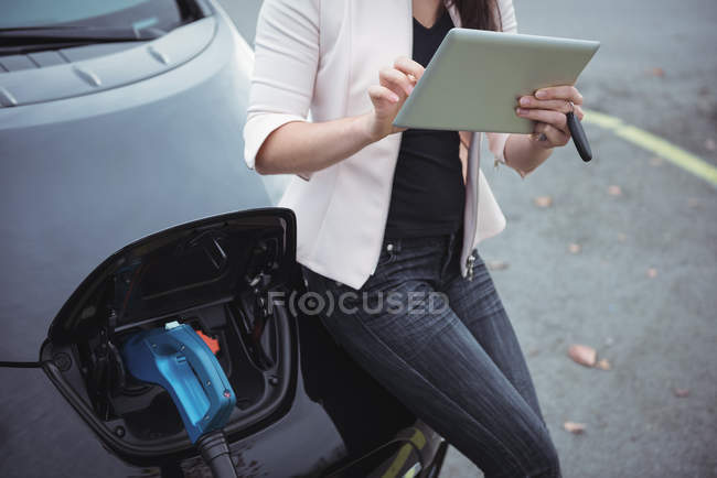 Средняя часть женщины с помощью цифрового планшета во время зарядки электромобиля на улице — стоковое фото