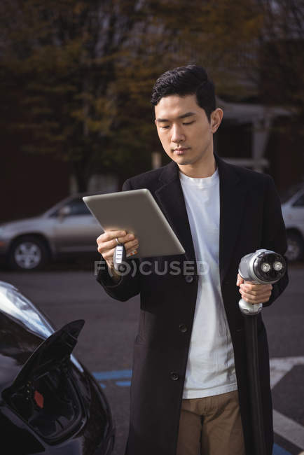 Uomo che utilizza tablet digitale mentre tiene il caricabatterie per auto presso la stazione di ricarica del veicolo elettrico — Foto stock
