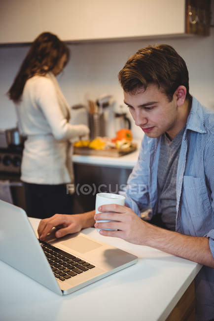 Homme utilisant un ordinateur portable tandis que la femme travaillant en arrière-plan à la cuisine — Photo de stock