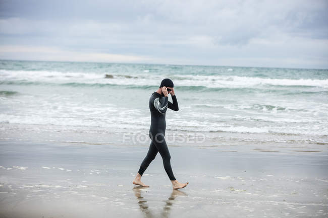 Athlete in wet suit wearing swimming goggles while walking on the beach — Stock Photo