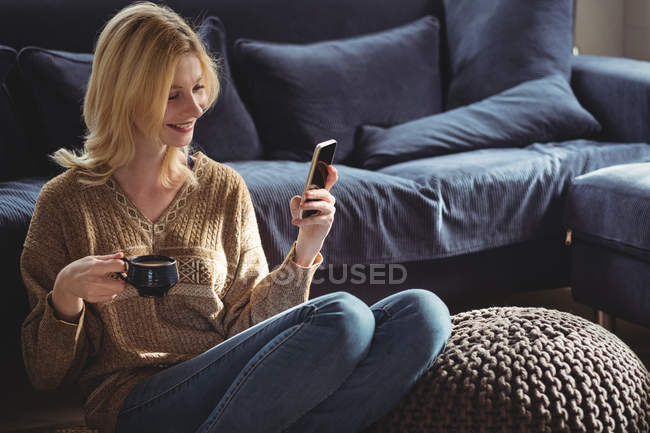 Belle femme utilisant un téléphone portable tout en prenant le thé dans le salon à la maison — Photo de stock