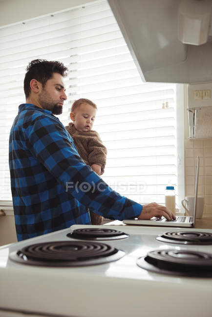Средний взрослый мужчина использует ноутбук, держа маленького сына дома на кухне — стоковое фото