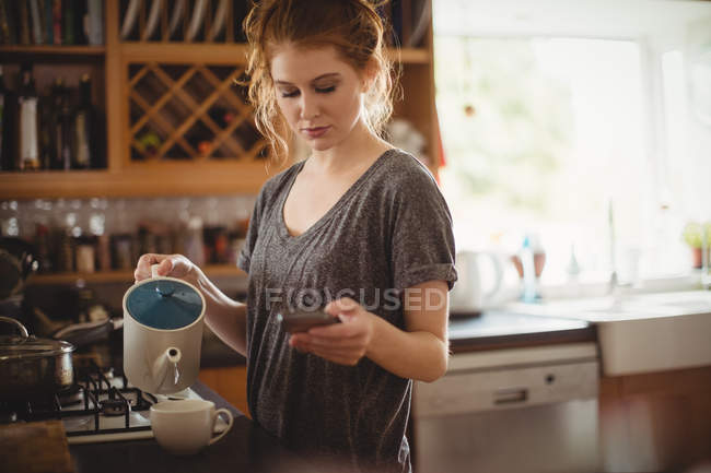 Красива жінка використовує мобільний телефон під час приготування кави на кухні вдома — стокове фото