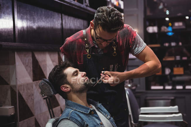 Man getting beard trimmed by barber with scissors in barber shop — Stock Photo