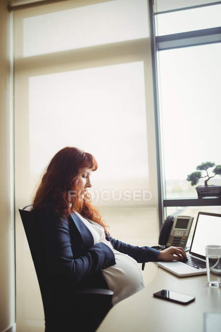 Grávida empresária tocando barriga ao usar laptop no escritório — Fotografia de Stock