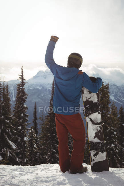 Задний вид человека с поднятой рукой, держащего сноуборд на горе против неба зимой — стоковое фото