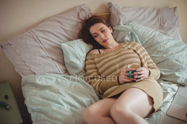 Thoughtful woman holding coffee cup and lying on bed at home — Stock Photo