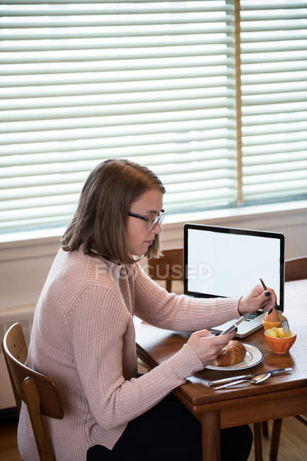 Donna che usa il telefono cellulare mentre fa colazione in soggiorno a casa — Foto stock