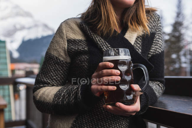 Средняя секция женщины в зимней одежде держит бокал пива на открытой террасе бара — стоковое фото