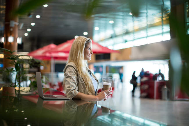 Улыбающаяся женщина с кофе, стоящая в зоне ожидания в аэропорту — стоковое фото