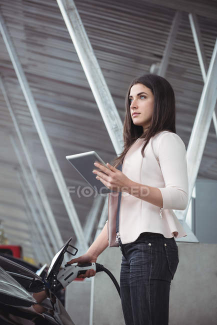 Beautiful woman using digital tablet while charging electric car at vehicle charging station — Stock Photo