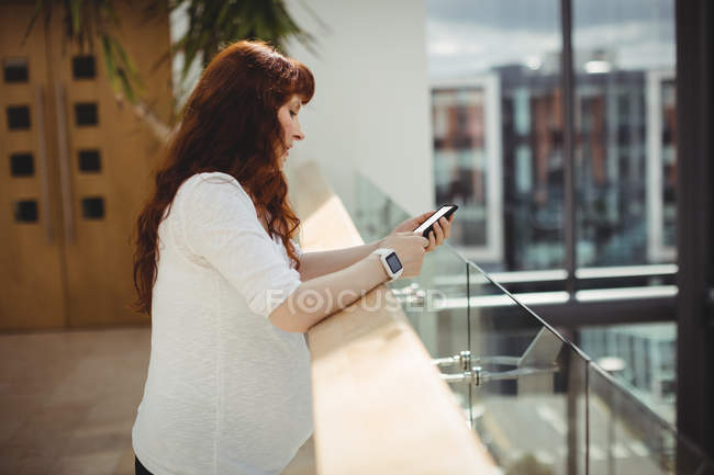 Grávida empresária usando telefone celular perto do corredor no escritório — Fotografia de Stock