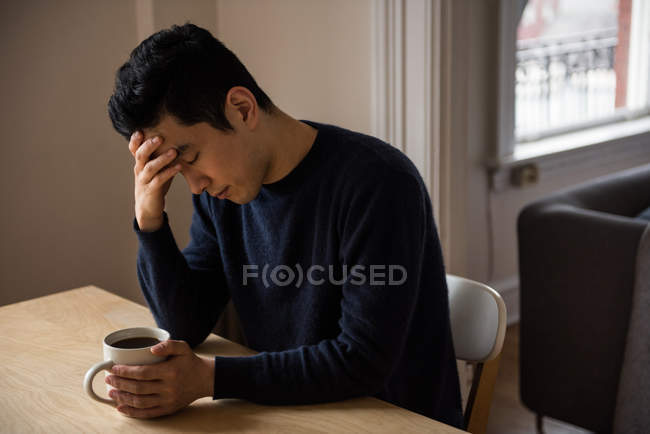 Hombre tenso sentado en la mesa con taza de café en casa - foto de stock