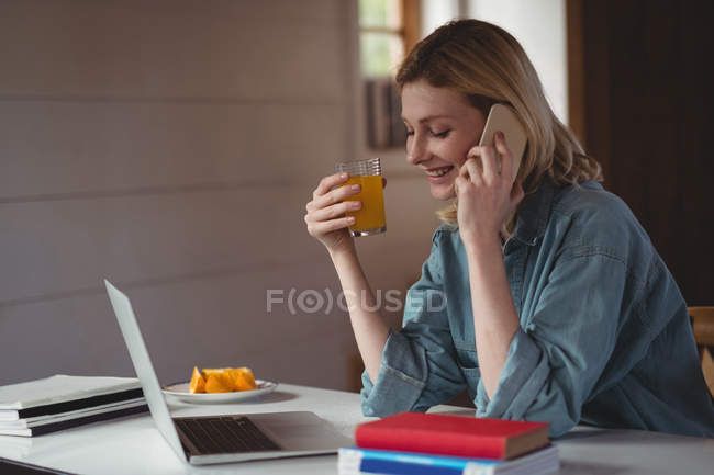 Красива жінка розмовляє на мобільному телефоні, маючи сік вдома — стокове фото