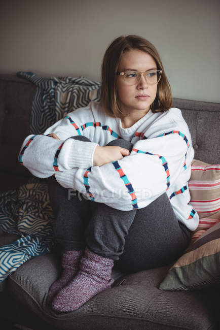 Mujer pensativa sentada y abrazando rodillas en la sala de estar en casa - foto de stock