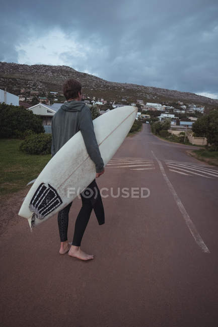 Low section of man carrying surfboard and shoes walking on road — Stock Photo