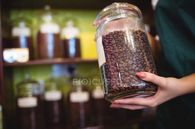 Sección media del tendero femenino sosteniendo frasco de granos de café en el mostrador en la tienda - foto de stock