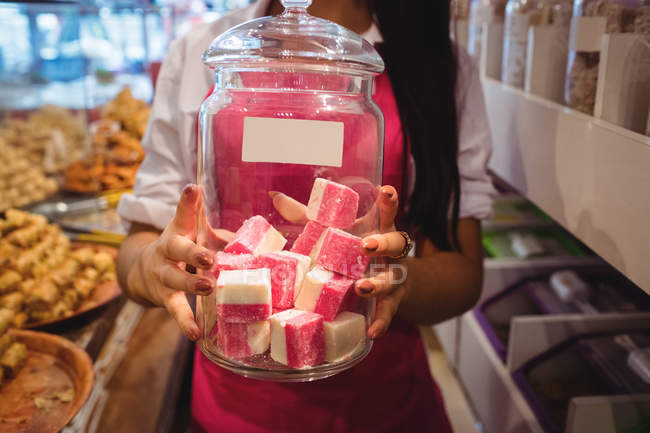 Sección media del tendero femenino sosteniendo tarro de dulces turcos en el mostrador en la tienda - foto de stock