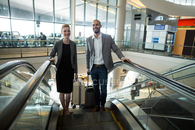 Empresários sorridentes com bagagem subindo na escada rolante no terminal do aeroporto — Fotografia de Stock