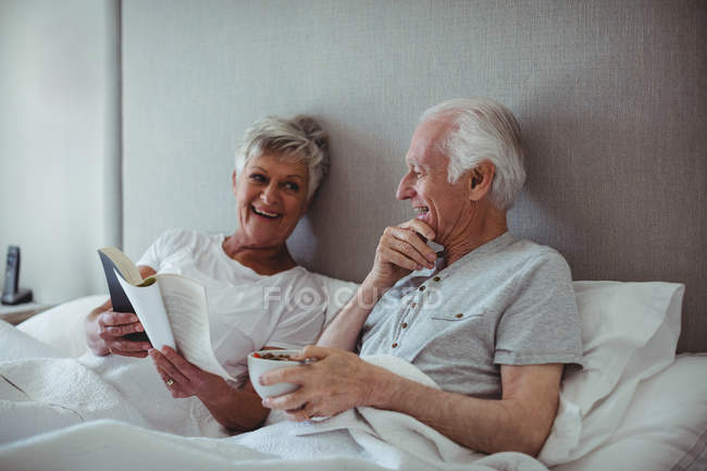 Senior man having breakfast while woman reading a book on bed in bed room — Stock Photo