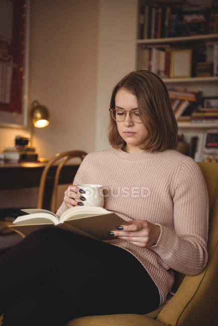 Mulher lendo livro enquanto toma café na sala de estar em casa — Fotografia de Stock