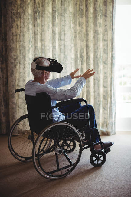 Пенсионер сидит на инвалидной коляске и пользуется гарнитурой виртуальной реальности в спальне дома — стоковое фото