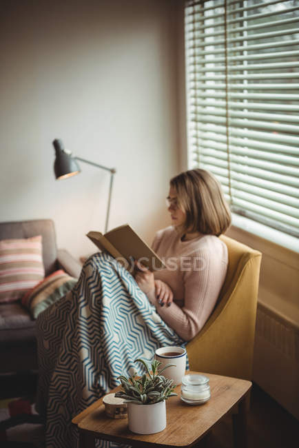 Mujer sentada en silla y leyendo libro en casa - foto de stock