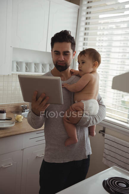 Hombre adulto medio usando tableta digital mientras sostiene al bebé en la cocina - foto de stock