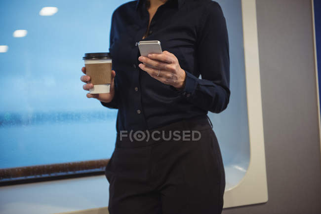 Середній розділ бізнес-леді з чашкою кави за допомогою телефону, стоячи в поїзді — стокове фото