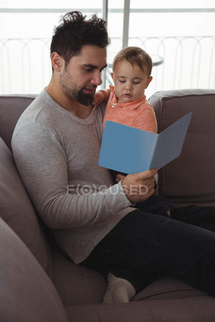 Vater liest Buch, während er sein Baby zu Hause hält — Stockfoto