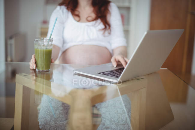 Средняя часть беременной женщины, использующей ноутбук во время употребления сока дома — стоковое фото