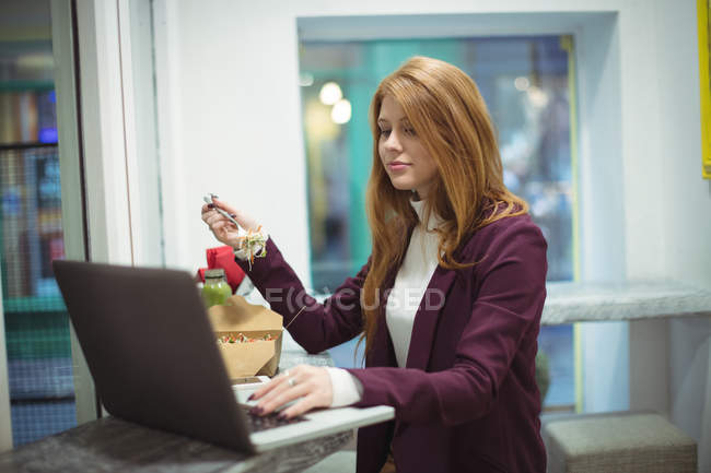Femme rousse utilisant un ordinateur portable tout en mangeant de la salade — Photo de stock
