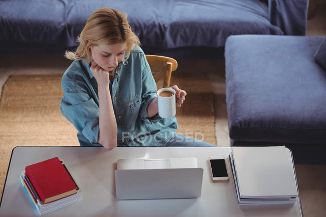 Красива жінка, що має каву, використовуючи ноутбук у вітальні вдома — стокове фото
