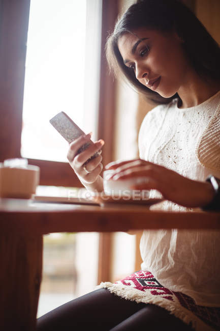 Hermosa mujer usando el teléfono móvil mientras toma una taza de café en la cafetería - foto de stock